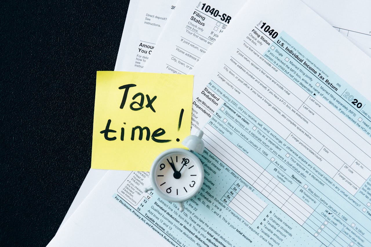 A stack of tax forms with a clock and yellow sticky note saying Tax time! indicating urgency.